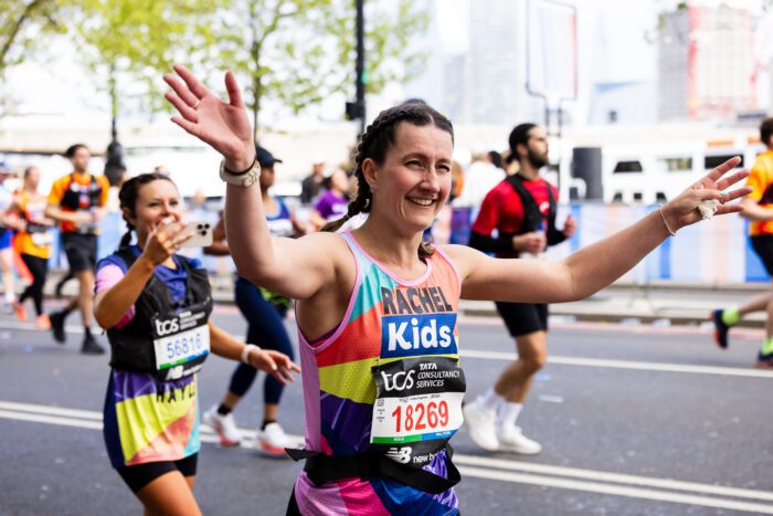 A woman is running with both hands up in the air. She is wearing a Kids vest. There are other runners behind her