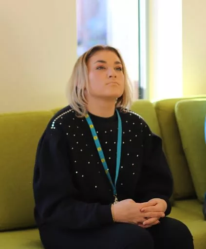 Georgia is sitting on a couch. She is wearing a black jumper and blue lanyard. She has short blond hair.