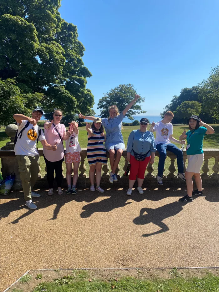 Young people are posing outside, the blue sky behind them. They are giving thumbs ups and lifting their arms in the air.